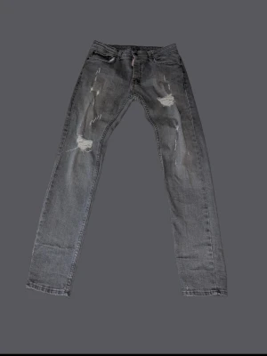 Grå slitna jeans från Dsquared2 - Säljer ett par grå jeans från Dsquared2 med snygga slitningar och trasiga detaljer på framsidan. Modellen har klassisk femficksdesign och raka ben. Jeansen är tillverkade i bomull och har en cool, urban vibe som passar perfekt till streetwear.