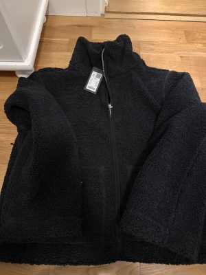 Svart teddyjacka stl S - Mysig svart teddyjacka  i storlek S. Jackan har hög krage, hel dragkedja framtill och stora fickor. Materialet är fluffigt och ger en skön vibe, perfekt för kyliga dagar. Enkel och stilren design som funkar till många outfits.