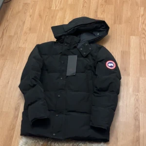 Svart dunjacka från Canada Goose - Säljer en svart dunjacka från Canada Goose med huva och avtagbar pälskant. Jackan har lång ärm, klassisk logga på armen och dragkedja med knappar framtill. Perfekt för kalla vinterdagar och riktigt snygg streetstil.