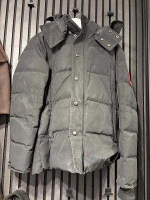 Svart pufferjacka från Canada Goose - Svart pufferjacka från Canada Goose med avtagbar huva och klassisk logotyp på ärmen. Jackan har knappar framtill, långärmade ärmar och är tillverkad i slitstark polyester. Perfekt för kalla vinterdagar och har en clean, oversized look.