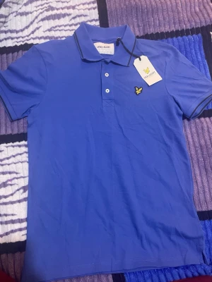 Blå pikétröja från Lyle & Scott - Säljer en fräsch blå pikétröja från Lyle & Scott i storlek S. Tröjan har klassisk krage, tre vita knappar och en gul broderad fågel på bröstet. Materialet är ekologisk bomull med lite elastan för extra komfort. Mörkblå kantdetaljer på kragen och ärmarna.