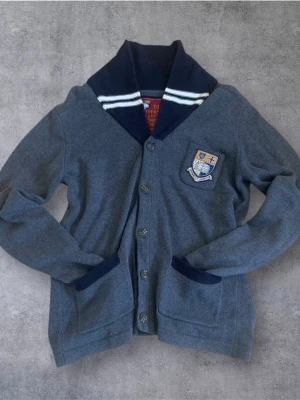 Vintage Morris Kofta - Snygg vintage kofta från Morris University Collection med marinblå krage och detaljer. Broderat emblem på bröstet, två fickor framtill och knappar hela vägen. Perfekt för dig som gillar klassisk och preppy stil. Inga tecken på slitage eller annan användning!!