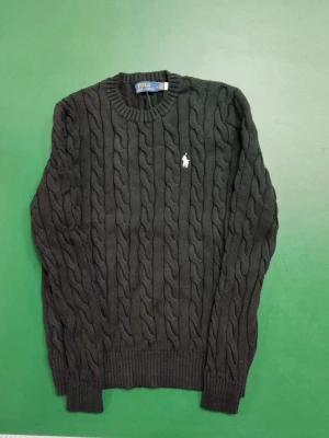 Svart stickad tröja Polo Ralph Lauren - Svart kabelstickad tröja från Polo Ralph Lauren i storlek M. Tröjan har rund hals, ribbade muddar och ett broderat vitt Polo-logo på bröstet. Perfekt för dig som gillar klassisk stil med en modern touch.