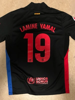 Barcelona fotbolls tröja Lamine Yamal#19 - Bilderna visar en svart FC Barcelona fotbollströja med röda och blå detaljer, Lamine Yamal och nummer 19 på ryggen, samt sponsorer som Spotify och UNHCR. Ingen sko syns på bilderna.