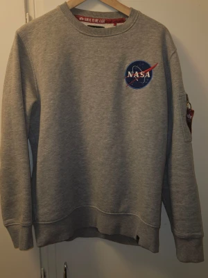 Grå NASA sweatshirt från Alpha Industries - Grå sweatshirt med NASA-logga framtill och stort Space Shuttle-tryck på ryggen. Har amerikansk flagga på ärmen och dragkedja med 'Remove Before Flight'-tag. Rund halsringning och ribbade muddar. Perfekt för dig som gillar rymden och coola detaljer.