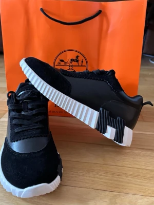 Svarta H3rm3s skor - Säljer ett par svarta H3rm3s sneakers i storlek 40. Skorna har en mix av mocka och skinn, med coola vita och svarta räfflade sulor och snörning. Snygg kontrast mellan material och detaljer, perfekt för dig som gillar exklusiva och unika sneakers.