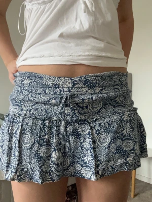 Blommig blåvit kortkjol med snörning - Supersöt kortkjol i blått och vitt med blommigt mönster och volangkant. Kjolen har en bred midja med snörning framtill som ger en chill vibe. Perfekt för varma dagar och enkel att matcha med en enkel topp. Materialet känns lätt och luftigt. Inbyggda shorts!!