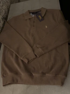 Brun tröja Polo Ralph Lauren - Säljer en brun långärmad pikétröja från Polo Ralph Lauren med klassisk krage och knappslå. Tröjan har det ikoniska broderade logot på bröstet och ribbade muddar vid ärmslut och nederkant. Perfekt för en chill och stilren look.