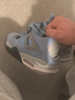 Nike Air Jordan 4 University Blue - Säljer ett par Nike Air Jordan 4 University Blue sneakers med ljusblå mocka, grå detaljer med svarta stänk och Jumpman-logga på hälen. Skorna har snörning, meshpaneler och en platt sula. Perfekt för dig som vill ha en unik och snygg sneaker med ikonisk design.