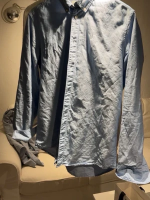 Ljusblå skjorta från East West - Snygg ljusblå skjorta från East West i storlek M. Skjortan har klassisk krage, knappar framtill och långa ärmar. Tillverkad i mjuk bomull som känns skön mot huden. Perfekt för dig som gillar stilrena och enkla plagg.