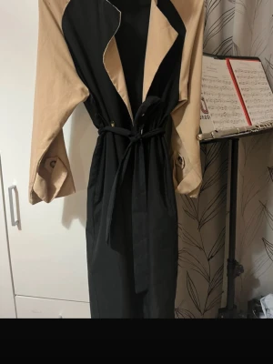 Svart och beige trenchcoat med bälte - Snygg lång trenchcoat i svart och beige med klassisk krage och knappar framtill. Jackan har ett bälte i midjan för att kunna knyta och justera passformen. Ärmarna är beige och har knappdetaljer vid mudden. Perfekt för dig som vill ha en stilren och cool look.