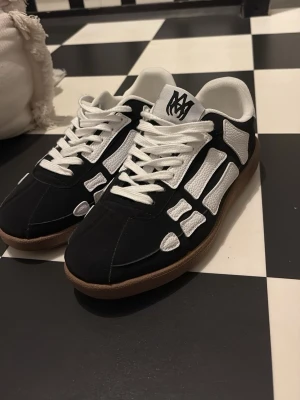 Svarta och vita sneakers från Amiri - Säljer ett par snygga Amiri sneakers i svart och vitt med coolt skelettmönster på sidorna. Skorna har vit snörning, bruna gummisulor och detaljer i skinn. Amiri-logga på hälen och plösen. Perfekt för dig som gillar streetwear och vill sticka ut.