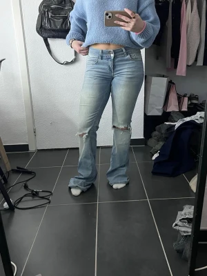 Ljusblå bootcut jeans från Nelly  - Säljer ett par ljusblå jeans med bootcut-modell och slitna hål på båda knäna. Jeansen har klassisk femficksdesign, låg midja och är tillverkade i denim. Perfekta för dig som gillar en avslappnad och trendig look.