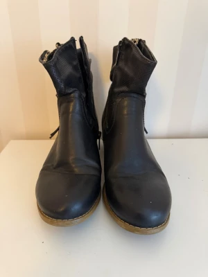 Svarta boots med leopardfoder och dragkedja - Säljer ett par svarta boots i skinn med perforerad ovandel och snygg dragkedja i guld på sidan. Insidan har ett coolt leopardmönstrat foder som syns när skaftet viks ner. Bootsens klack är låg och i trälook, och bak finns en dekorativ knappdetalj.