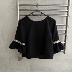 Svart blus med volangärm och guld detalj  - Säljer en svart blus med rund hals och volangärmar. Ärmarna har en snygg detalj guld detalj som ger extra glans. Blusen är lite croppad och har en knapp i nacken för enkel påklädning. Perfekt för dig som gillar stilrena plagg med lite extra edge.