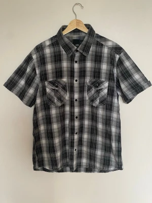 Svartvit rutig skjorta Jack & Jones - Snygg kortärmad skjorta från Jack & Jones i svartvitt rutigt mönster. Skjortan har två bröstfickor med lock och knappar framtill. På ryggen finns broderad text 'Huntington' och detaljerad brodyr. Perfekt för dig som gillar streetstyle.