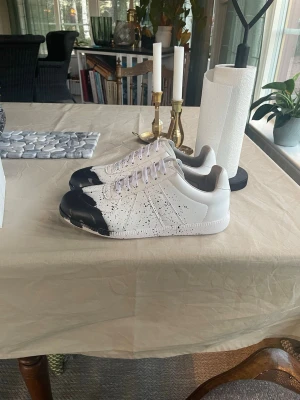 Maison Margiela - Maison Margiela sneakers i vitt skinn med svart färg på tån och svarta färgstänk över hela skon. Klassisk låg modell med snörning och vit sula. Insidan är fodrad i beige och skon har en clean, minimalistisk look med en konstnärlig twist.