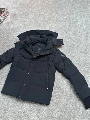 Svart dunjacka från Canada Goose - Säljer en svart dunjacka från Canada Goose med avtagbar huva och diskret logga på ärmen. Jackan har dragkedja och knappar framtill, samt ribbade muddar vid ärmsluten. Perfekt för kalla vinterdagar och riktigt snygg till alla outfits. 