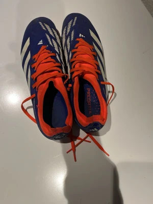 Adidas Predator fotbollsskor - Säljer ett par Adidas Predator fotbollsskor i blått med vita detaljer och orange snörning. Skorna har en låg profil och är designade för gräsplan, med mönstrad ovandel och klassiska Adidas-ränder på sidan. Insidan är också orange för extra färgklick. Storlek 42 men de sitter som storlek 40-41.