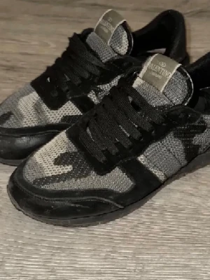 Valentino Garavani sneakers med camomönster - Snygga sneakers från Valentino Garavani med svart och grått camouflagemönster. Skorna har snörning, rund tå och platt sula. Ovandelen är i mesh och mocka, vilket ger en cool och modern vibe. Perfekta för dig som gillar streetstyle och exklusiva märken.