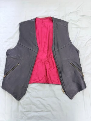 Svart skinnväst från Bondelid - Cool svart skinnväst från Bondelid med starkt rosa foder. Västen har två sneda fickor med dragkedja fram och ett justerbart bälte bak. Obs kedjan framtill är trasig!