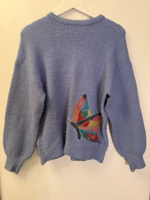 Ljuslila stickad tröja med fjäril - Mysig ljuslila stickad tröja med rund hals och ribbade muddar. På ena sidan finns en stor, färgglad fjäril i blått, rött, gult och grönt som ger en unik touch. Perfekt för dig som gillar kreativa detaljer och vill sticka ut.
