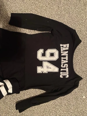 Svart kortklänning med tryck och ränder - Cool svart klänning med vita ränder längst ner och trycket 'FANTASTIC 94' på magen. Klänningen har trekvartsärm och en sportig look med rund hals. Materialet känns mjukt och stretchigt, perfekt för dig som gillar en avslappnad stil. För barn storlek 134/140