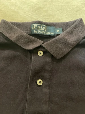 Svart pikétröja från Polo Ralph Lauren - Klassisk mörkblu pikétröja från Polo Ralph Lauren i storlek M. Tröjan har korta ärmar, krage och två knappar framtill. På bröstet finns det ikoniska röda broderade Polo-logot. Materialet är mjuk bomull som känns skönt mot huden.