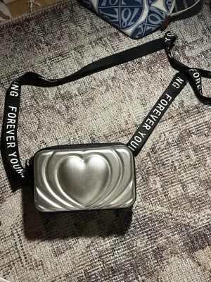 Silver axelväska med hjärtdetalj - Säljer en cool axelväska i silver med ett stort hjärta på framsidan. Väskan har svart justerbar rem med texten 'FOREVER YOUNG' och dragkedja. Insidan har flera fack i mesh för småsaker. Pris kan diskuteras!!💕