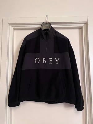 OBEY Svart fleecejacka  - Svart fleecejacka från OBEY med vit logotyp broderad över bröstet. Jackan har half zip och hög krage med snygg beige detalj. Loose oversize passform och känguru-ficka. Jättesnygg streetstyle fleece som håller varm. 2950kr originalpris. Mycket bra skick, som ny! Säljs på flera sidor, först till kvarn. 