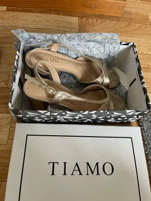 Guldiga sandaler med klack från Tiamo - Säljer ett par snygga klackar från Tiamo i  guld med bred klack och öppen tå. Skorna har ett stilrent korsat band över foten och justerbart spänne vid ankeln. Perfekta för dig som vill ha en chic look med lite höjd❤️Köpte dom för 699 men säljer för 400. (endast använt en gång vid min bal!)