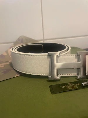 115cm - Bilderna visar ett vitt skärp med en stor H-formad metallspänne, inte en väst. Skärpet verkar vara gjort av skinn och har tydliga sömmar längs kanterna.