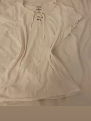 Vit t-shirt med snörning från SMOG - Säljer en vit t-shirt från SMOG i storlek XS. T-shirten har korta ärmar och cool snörning vid halsen som ger en extra detalj. Materialet är mjuk bomull och passformen är normal. Perfekt för dig som gillar enkel stil med en twist.