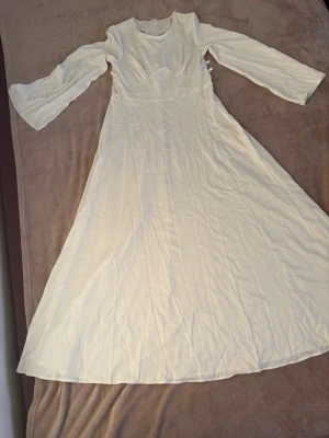 Vit långklänning med långa ärmar - Säljer en elegant vit långklänning med rund halsringning och långa, lätt utsvängda ärmar. Klänningen har en markerad midja och är tillverkad i ett mjukt, slätt material som ger ett fint fall. Dragkedja i ryggen för enkel påklädning.