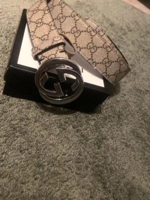 Gucci beige GG-mönstrad bälte - Säljer ett snyggt Gucci-bälte i beige med klassiskt GG-mönster och en stor rund metallspänne med logga. Bältet har en lyxig känsla och är tillverkat i slitstarkt material med läder på insidan. Perfekt för att lyfta din outfit med en exklusiv touch.