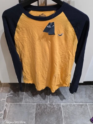 Gul och marinblå långärmad topp Hollister - Säljer en långärmad topp från Hollister i storlek S. Toppen har gul kropp och marinblå ärmar samt krage, med broderad Hollister-logga på bröstet. Materialet är mjuk bomull och modellen har en rund hals. Perfekt för dig som gillar sportig och avslappnad stil.