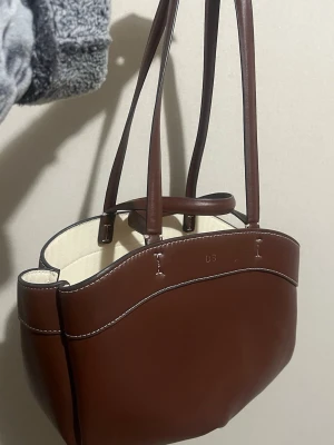 Brun axelväska från ZARA med initialer ”D.S” - Snygg brun axelväska från Zara med stilrena sömmar och ljus insida. Väskan har långa handtag och ett innerfack med dragkedja i beige tyg. Perfekt storlek för att få plats med allt du behöver och passar till många olika looks. Initialer ”D.S”