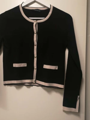 Svart kofta med pärlknappar från Zara - Snygg svart kofta från Zara med beige kantband och dekorativa pärlknappar framtill. Koftan har två fejkade fickor och är långärmad med beige detaljer vid ärmslut och nederkant. Stickad i mjukt material, perfekt för lager-på-lager under hösten.