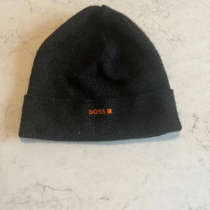 Svart mössa från HUGO BOSS - Svart mössa från HUGO BOSS med orange broderad logga framtill. Klassisk form med uppvikt kant, perfekt för kalla dagar. Materialet är mjukt och stickat för extra komfort.