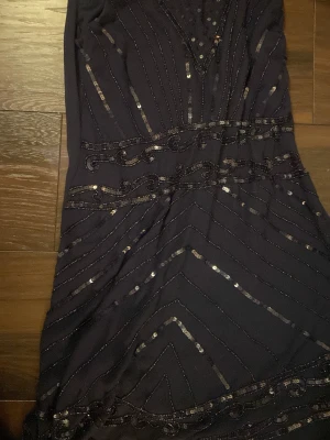 Mörkblå glittrig långklänning Anna Field - Snygg mörkblå långklänning från Anna Field med glittriga paljettdetaljer och dekorativa mönster. Klänningen har v-ringad hals och är figurnära med elegant fall. Perfekt för dig som vill sticka ut med stil och glitter.