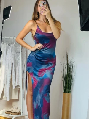 Lång mönstrad klänning med slits ny med prislapp - Säljer en lång klänning från Micas med tunna axelband och snygg slits eller utskärning på sidan. Klänningen har ett färgstarkt mönster i blått och vinrött med inslag av lila och rosa. Materialet är glansigt och känns lyxigt mot huden. Perfekt för dig som vill sticka ut! Ny med etiketter kvar. Storlek L men väldigt stretchig, armhåla till armhåla 42 cm, längd 145 cm