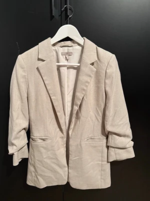 Beige kavaj med trekvartsärm - Snygg beige kavaj med klassisk krage och trekvartsärmar. Jackan har två fickor framtill och är tillverkad i ett mjukt material som ger en stilren look. Perfekt för dig som vill ha en enkel men trendig kavaj i garderoben.