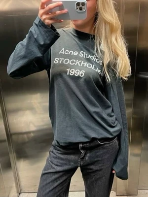 Distressed Mörkgrå/Navy långärmad t-shirt Acne Studios Stockholm 1996 - Säljer en mörkgrå långärmad t-shirt från Acne Studios med vit text 'Acne Studios Stockholm 1996' på bröstet. Unisex-modell med rund hals och relaxed fit. Materialet är mjuk bomull som känns skönt mot huden. Perfekt för dig som gillar stilrena och coola plagg.