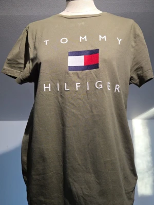 Olivgrön Tommy Hilfiger t-shirt - Snygg olivgrön t-shirt från Tommy Hilfiger med stort broderat logotyptryck i vitt, rött och marinblått på bröstet. Klassisk rund hals och korta ärmar. Tillverkad i mjuk bomull som är skön mot huden. Perfekt för dig som gillar stilrena märkesplagg.