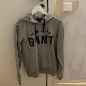 Grå hoodie från GANT med tryck - Säljer en grå hoodie från GANT med svart tryck 'NEW HAVEN GANT' på bröstet. Tröjan har huva med svarta snören och ribbade muddar vid ärmslut och nederkant. Perfekt för en avslappnad och sportig stil.