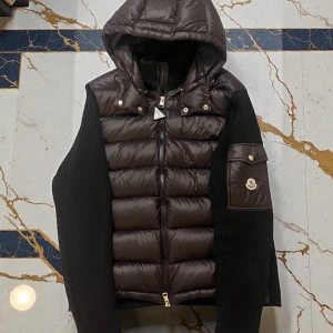 Svart cardigan med huva från Moncler med kvitto - Säljer en svart cardigan från Moncler med huva och ribbstickade ärmar. Västen har en quiltad framsida, dragkedja och en ficka med Moncler-logga på ärmen. Perfekt för dig som vill ha en snygg och varm look under höst och vinter. Tilkommer Kvitto!!