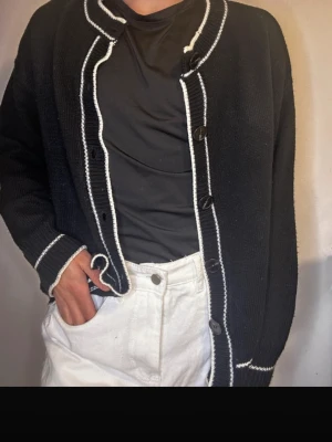 Svart kofta med vita detaljer - Snygg svart kofta med vita kantdetaljer och stora svarta knappar framtill. Koftan är stickad och har långärmade ärmar med vita ränder vid muddar och längs knappslån. Passar perfekt till jeans eller kjol för en chill och stilren look.