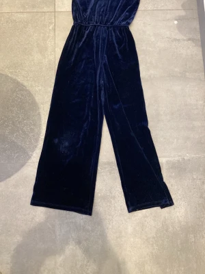 Mörkblå sammetsbyxdress från Kappahl - Snygg mörkblå byxdress i mjuk sammet från Kappahl, med puffärm och rund hals. Byxdressen har en öppen rygg med knappstängning och ett elastiskt midjeband för skön passform. Perfekt för dig som gillar att sticka ut med stil och komfort.