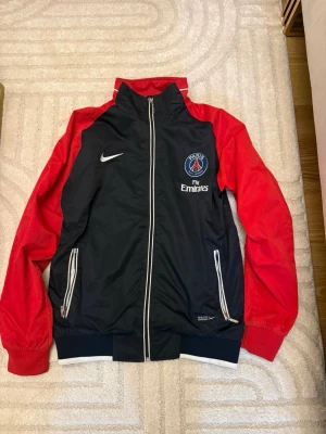 PSG vindjacka från Nike röd/blå - Säljer en snygg PSG vindjacka från Nike med röd färg på ärmarna och mörkblå kropp. Jackan har dragkedja framtill, två fickor med dragkedja och Paris Saint-Germain-logga på bröstet. Perfekt för dig som gillar fotboll och vill ha sportig stil.