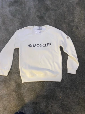 Vit sweatshirt från Moncler - Är lite små i storlek den är storlek M men säger mer som S eller XS.    Säljer en stilren vit sweatshirt från Moncler med svart broderad logga på bröstet. Tröjan har rund hals, långa ärmar och ribbade muddar vid ärmslut och nederkant. Perfekt för dig som gillar exklusiva märken och enkel design.